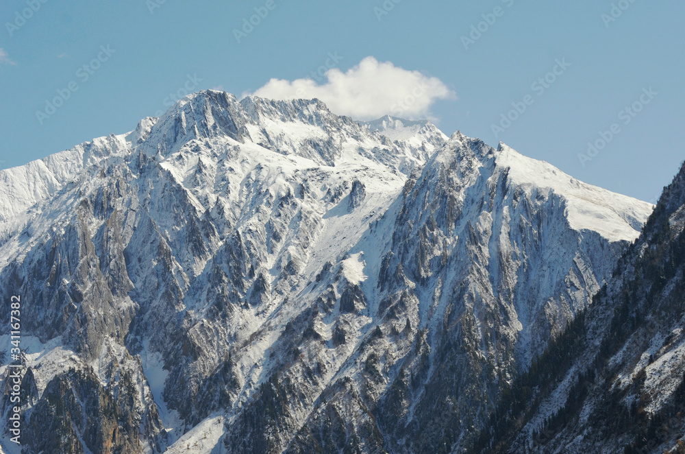 A mountain peak covered in snow rises into a blue sky. One white cloud hovers beside the peak. The sides of the mountain are covered in trees.
