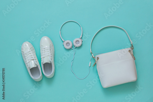 White women's bag, white headphones and white sneakers on a light blue background. Flat lay.