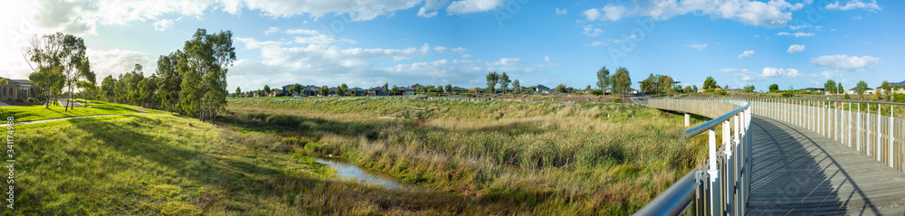 Fototapeta premium Panoramic view of Skeleton Waterholes Creek with a wooden boardwalk leads to some suburban houses in distance. Truganina, Melbourne, VIC Australia.