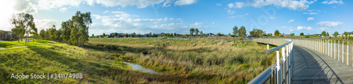 Panoramic view of Skeleton Waterholes Creek with a wooden boardwalk leads to some suburban houses in distance. Truganina, Melbourne, VIC Australia.