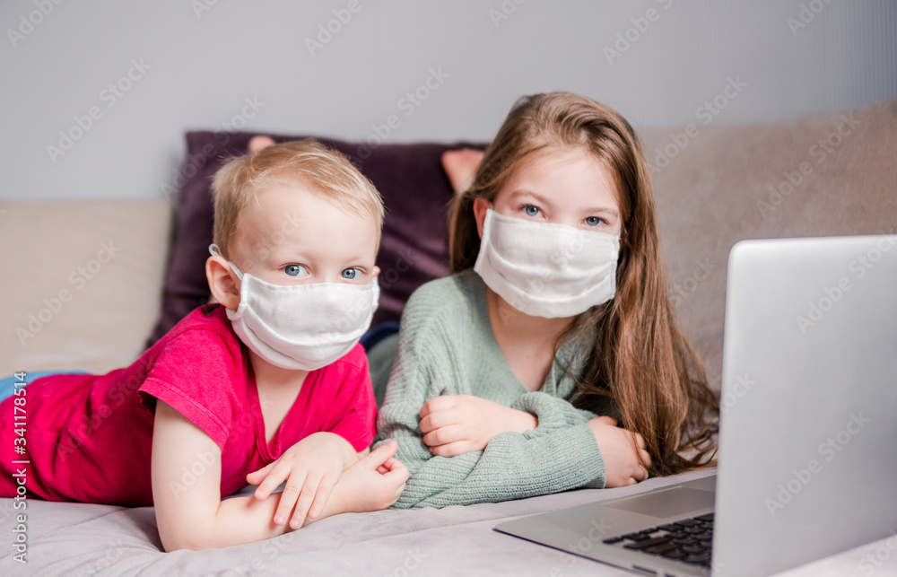 Two children are lying on a sofa at home in white medical masks and looking at the camera, a laptop next to the children. Concept of quarantine, self-isolation and epidemic Two children are lying on a sofa at home in white medical masks and looking at the camera, a laptop next to the children. Concept of quarantine, self-isolation and epidemic