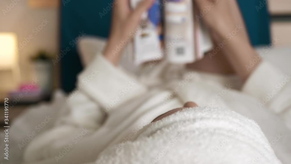 Girl reading magazine. Blurred view of woman in bedroom in white bathrobe lies on her back and turns over pages of fashion magazine. Close-up Girl reading magazine. Blurred view of woman in bedroom in white bathrobe lies on her back and turns over pages of fashion magazine. Close-up