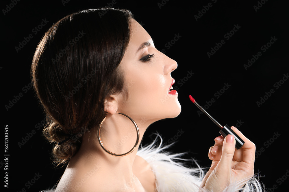 Obraz premium Beautiful young woman applying lipstick against dark background