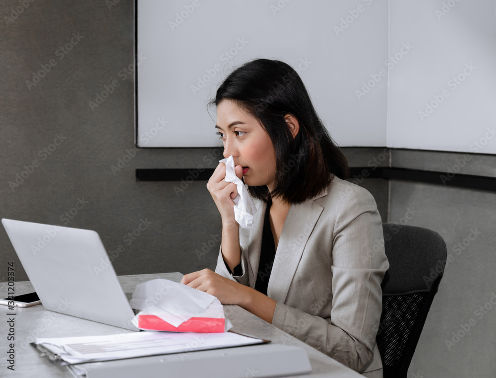 Sick bussinesswoman sitting at office desk with laptop, sneezing and coughing in facial tissue while working in meeting room. Working and influenze virus concept. Sick bussinesswoman sitting at office desk with laptop, sneezing and coughing in facial tissue while working in meeting room. Working and influenze virus concept.