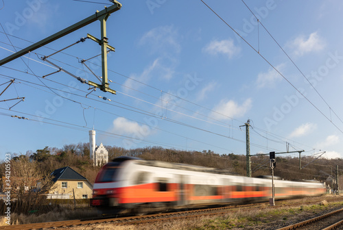 On a railway line on the German island of Rügen in the Baltic Sea. A regional train passes quickly. In the background the white castle of Lietzow on a mountain. Blue sky and clouds.