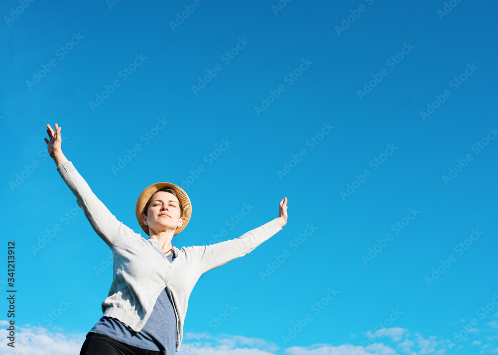 Happy woman wearing grey top, straw hat, raised her hands high against blu sky Happy woman wearing grey top, straw hat, raised her hands high against blu sky
