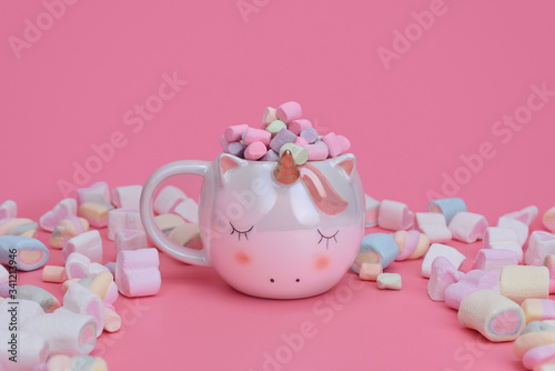 Sleeping unicorn cup on a pink background with mixed marshmallows and place for text. Sweets concept with copy space. Different multicolor marshmallows lie next to the unicorn.