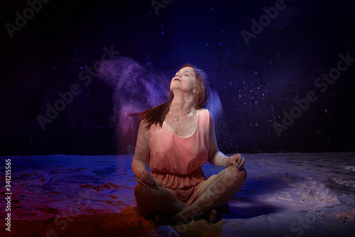 Beautiful woman with long brunette hair in pink dress with colored lights and flour around. Portrait of girl and female model posing in photoshoot with flour at studio with black background