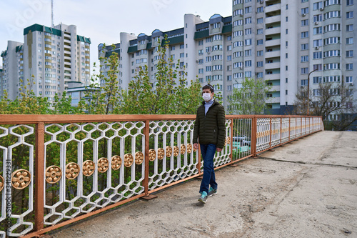 teen boy walks down the street during the day, a pedestrian walkway and high-rise buildings with apartments, a residential area, a medical mask on his face protects against viruses and dust