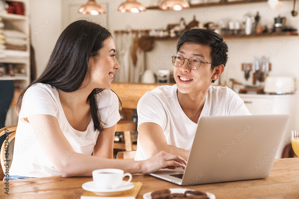 Image of happy couple smiling and using laptop while sitting at table Image of happy couple smiling and using laptop while sitting at table