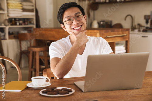 Image of asian man smiling and using laptop while sitting at table