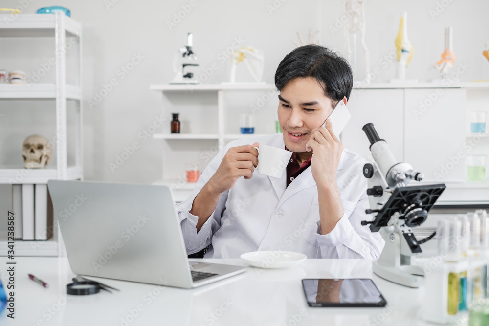 A male scientist with black hair wearing white coat using a mobile phone device sipping coffee with a laptop and a microscope in a laboratory setting with test tubes. Looking relax during break time. A male scientist with black hair wearing white coat using a mobile phone device sipping coffee with a laptop and a microscope in a laboratory setting with test tubes. Looking relax during break time.
