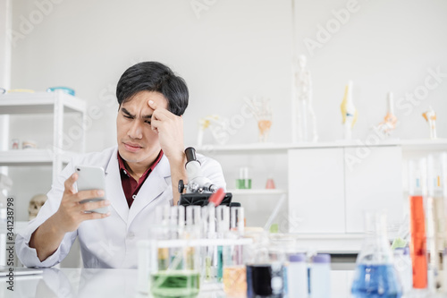 A young male scientist with black hair wearing white coat looking at a mobile phone device in his hand with another hand on the head. Looking tired and stressed in a laboratory setting.