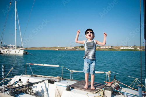 happy active small caucasian kid in sailor hat shouting with joy on white yacht board during summer travel vacation