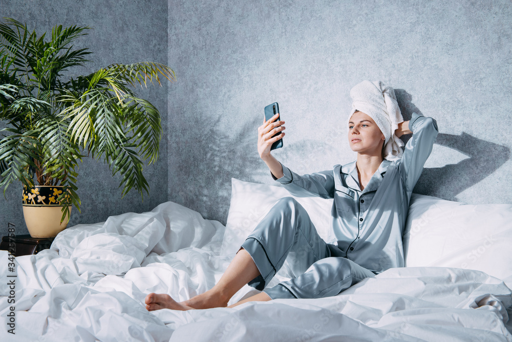 woman in pajamas and a towel on her head sits on a bed in the morning ...
