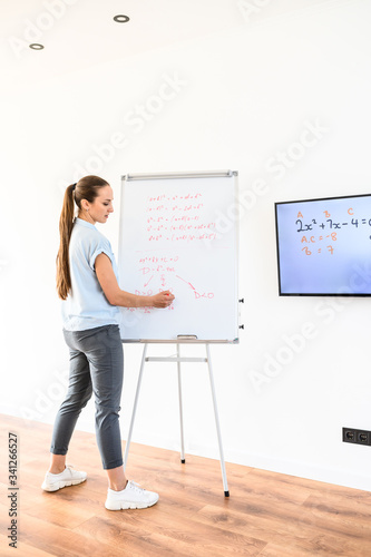 Young woman in smart casual writes formulas on the blackboard, some formulas on the screen on the wall. School teacher, schoolmaster
