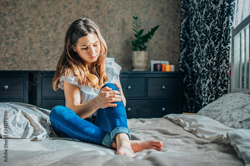 young brunette girl in a white blouse and blue jeans sits on a bed in her room, clutching a sore knee