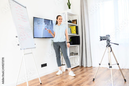 A woman is recording tutorial video at home, uses flip chart and monitor. Camera on tripod in front of woman. Online study concept
