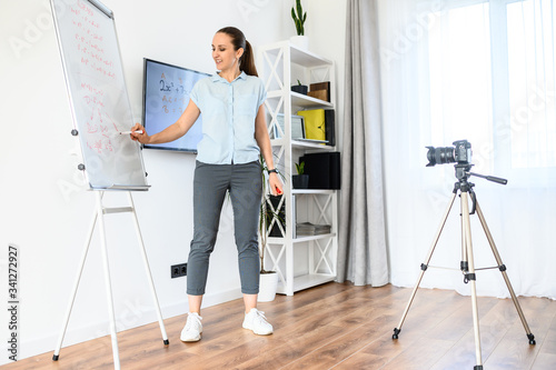 A woman is recording tutorial video at home, uses flip chart and monitor. Camera on tripod in front of woman. Online study concept