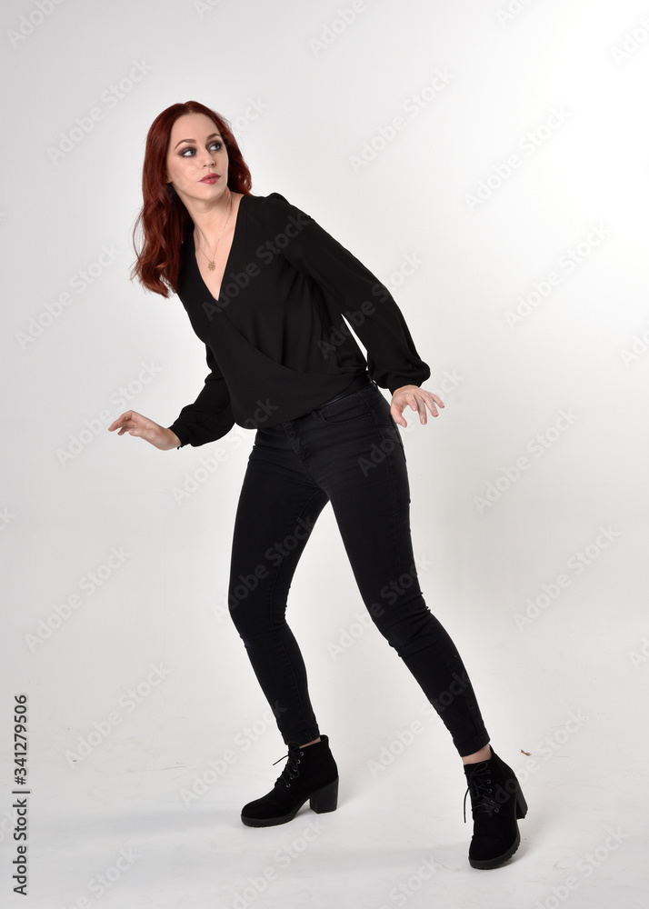 Portrait of a pretty girl with red hair wearing black jeans, boots and a blouse. full length standing pose, facing the camera, on a studio background. Portrait of a pretty girl with red hair wearing black jeans, boots and a blouse. full length standing pose, facing the camera, on a studio background.