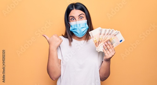 Young woman wearing protection mask for coronavirus holding 50 euros banknotes pointing thumb up to the side smiling happy with open mouth