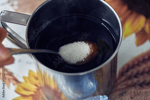 A teaspoon with sugar is lowered into an iron mug with tea, coffee. Close-up.