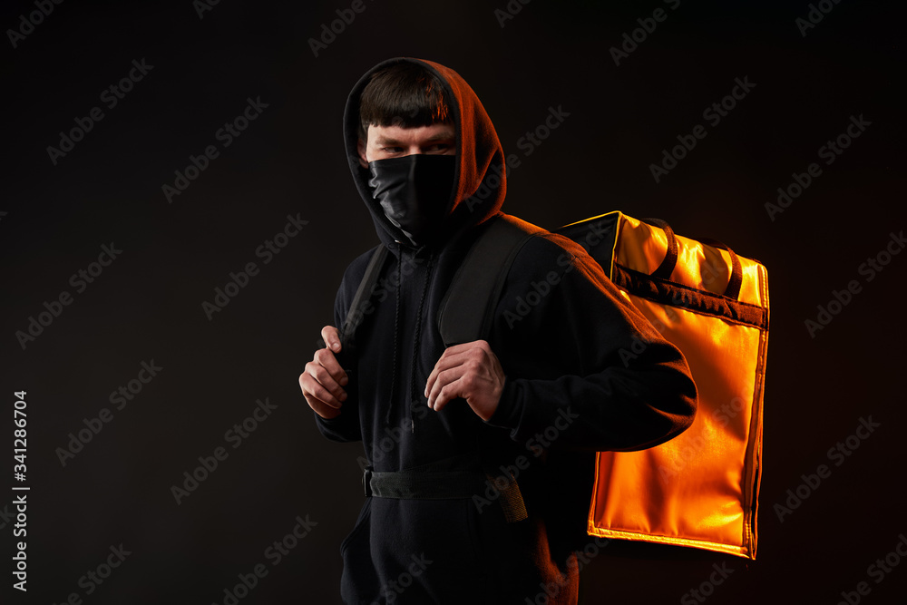 delivery man in black clothes in medicine mask with a yellow backpack on a black background with orange light