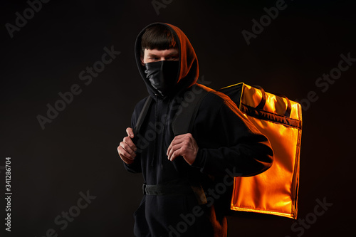 delivery man in black clothes in medicine mask with a yellow backpack on a black background with orange light