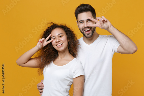 Cheerful young couple two friends european guy african american girl in white t-shirts posing isolated on yellow background studio. People lifestyle concept. Mock up copy space. Showing victory sign.