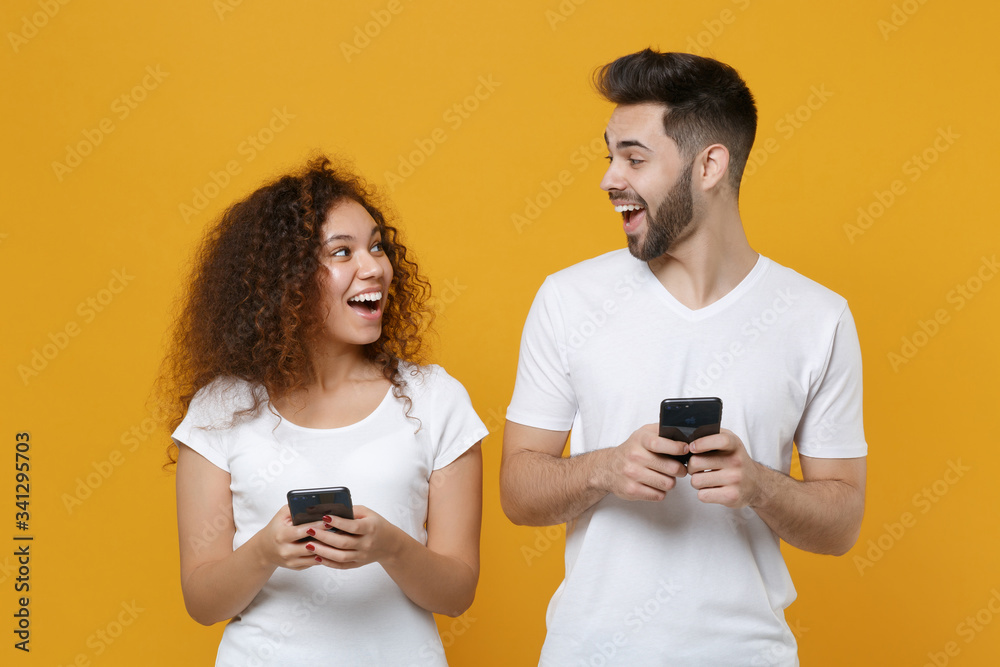 Excited young couple friends european guy african american girl in white t-shirts isolated on yellow background. People lifestyle concept. Using mobile phone typing sms message, looking at each other.