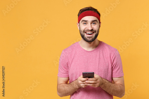 Cheerful young bearded fitness sporty guy sportsman in headband t-shirt in home gym isolated on yellow background. Workout sport motivation lifestyle concept. Using mobile phone, typing sms message. © ViDi Studio
