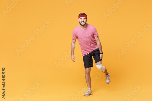 Ingured dissatisfied young bearded fitness guy 20s sportsman in headband t-shirt in home gym isolated on yellow background. Workout sport motivation concept. Touching leg with elastic bandage on knee. photo