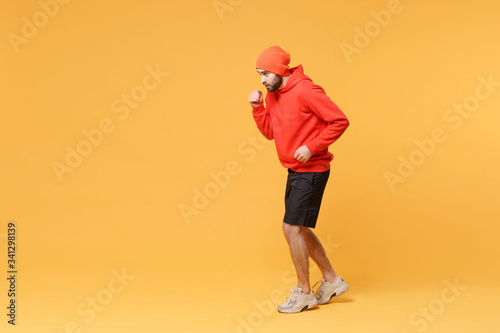 Side view of young bearded fitness sporty guy sportsman in hat, hoodie, shorts spend weekend in home gym isolated on yellow background in studio. Workout sport motivation lifestyle concept. Running.