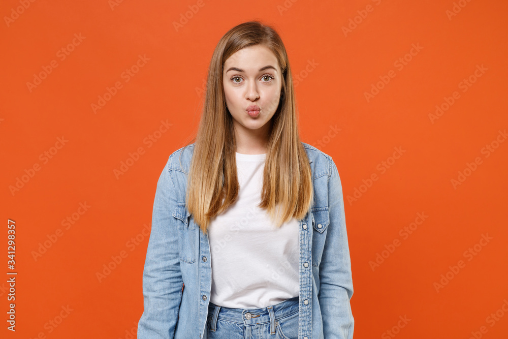 Fototapeta premium Pretty young woman girl in casual denim clothes posing isolated on bright orange wall background studio portrait. People sincere emotions lifestyle concept. Mock up copy space. Blowing cheeks, lips.