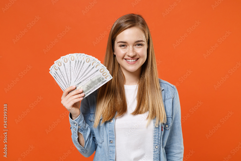 Smiling young woman girl in casual denim clothes posing isolated on orange wall background studio portrait. People lifestyle concept. Mock up copy space. Holding fan of cash money in dollar banknotes. Smiling young woman girl in casual denim clothes posing isolated on orange wall background studio portrait. People lifestyle concept. Mock up copy space. Holding fan of cash money in dollar banknotes.