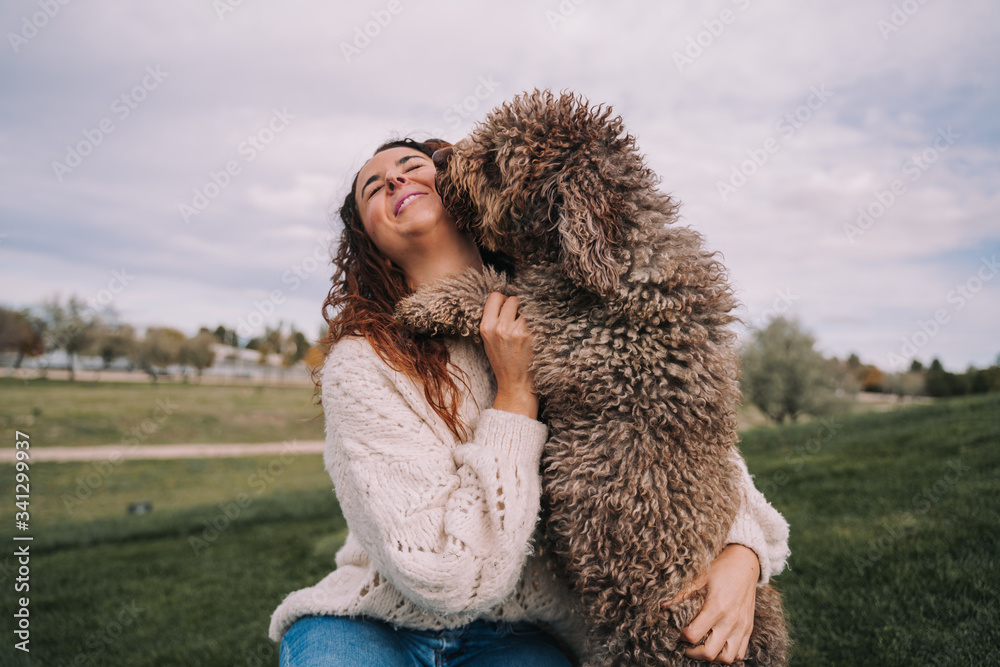 A beautiful woman is in the meadow with her dog. The owner is hugging her pet while the dog is licking her face.They are enjoying a day in the park. The pet is a Spanish water dog with brown fur. A beautiful woman is in the meadow with her dog. The owner is hugging her pet while the dog is licking her face.They are enjoying a day in the park. The pet is a Spanish water dog with brown fur.
