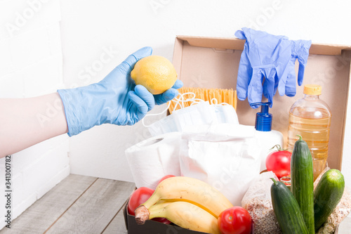 a woman in a rubber glove cakes a lemon in a box with food and hygiene products for a donation, volunteer work during the coronavirus pandemic, contactless delivery concept