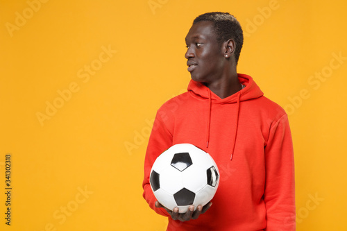 Handsome young african american man guy football player in red streetwear hoodie isolated on yellow wall background. Sport leisure lifestyle concept. Playing football hold soccer ball looking aside. © ViDi Studio