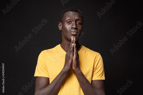 Pleading young african american man guy in yellow t-shirt posing isolated on black background studio portrait. People emotions  lifestyle concept. Mock up copy space. Holding hands folded in prayer.