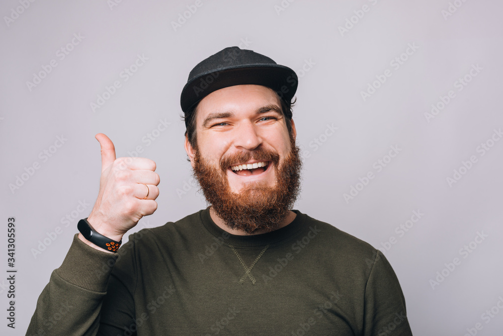 Photo of young happy man with beard showing thumbs up gesture over white wall Photo of young happy man with beard showing thumbs up gesture over white wall