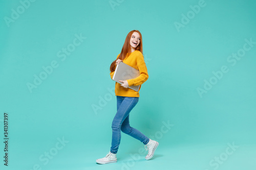 Laughing young redhead girl in yellow sweater posing isolated on blue turquoise wall background studio portrait. People lifestyle concept. Mock up copy space. Hold laptop pc computer, looking aside.