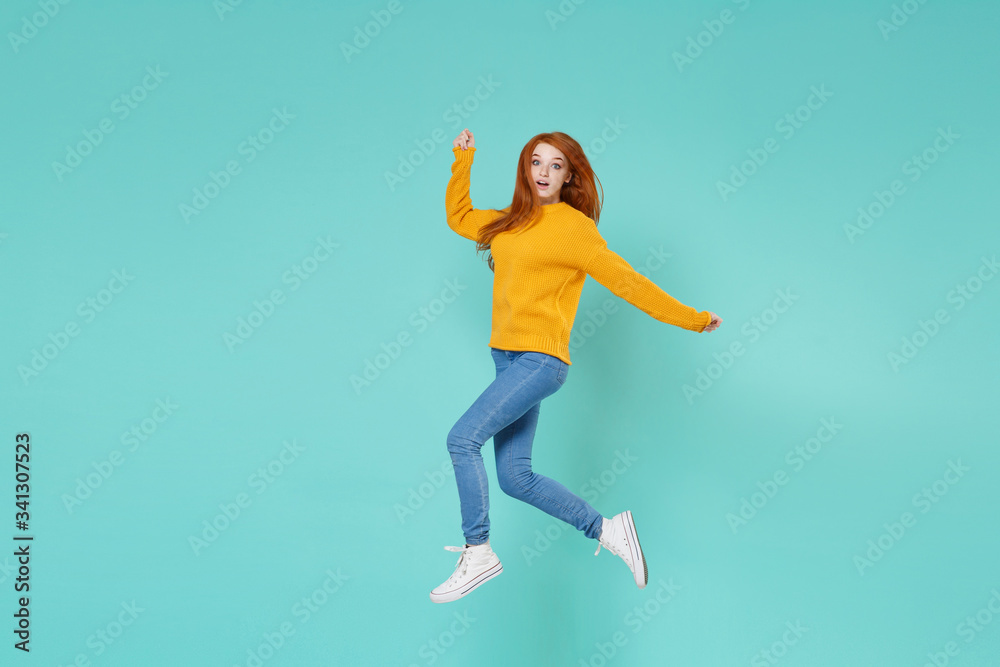 Shocked young redhead woman girl in yellow knitted sweater posing isolated on blue turquoise background studio portrait. People emotions lifestyle concept. Mock up copy space. Jumping spreading hands.