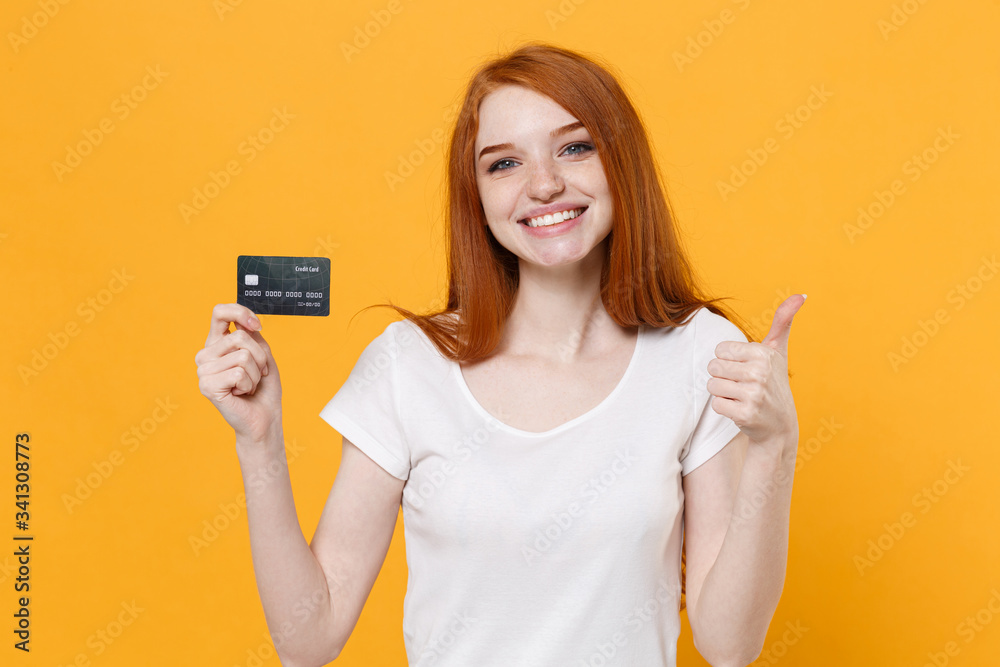 Smiling young redhead woman girl in white blank empty t-shirt posing isolated on yellow wall background in studio. People lifestyle concept. Mock up copy space. Hold credit bank card showing thumb up. Smiling young redhead woman girl in white blank empty t-shirt posing isolated on yellow wall background in studio. People lifestyle concept. Mock up copy space. Hold credit bank card showing thumb up.