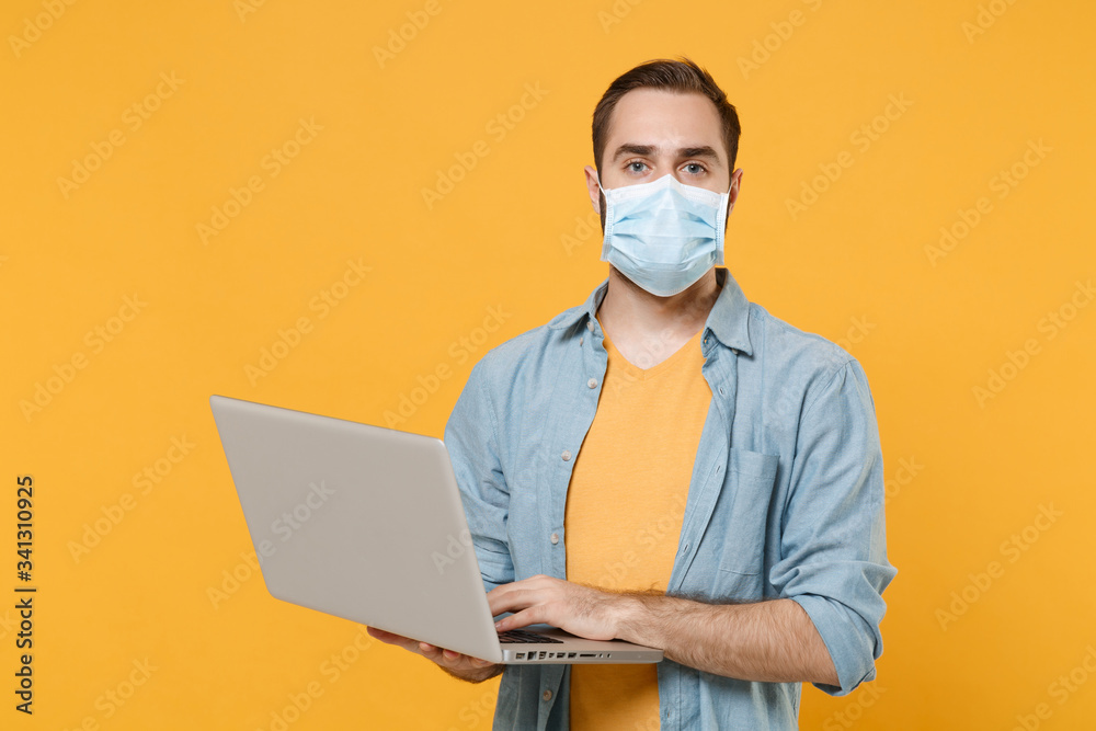 Young man in sterile face mask posing isolated on yellow background studio portrait. Epidemic pandemic spreading coronavirus 2019-ncov sars covid-19 flu virus concept. Working on laptop pc computer. Young man in sterile face mask posing isolated on yellow background studio portrait. Epidemic pandemic spreading coronavirus 2019-ncov sars covid-19 flu virus concept. Working on laptop pc computer.