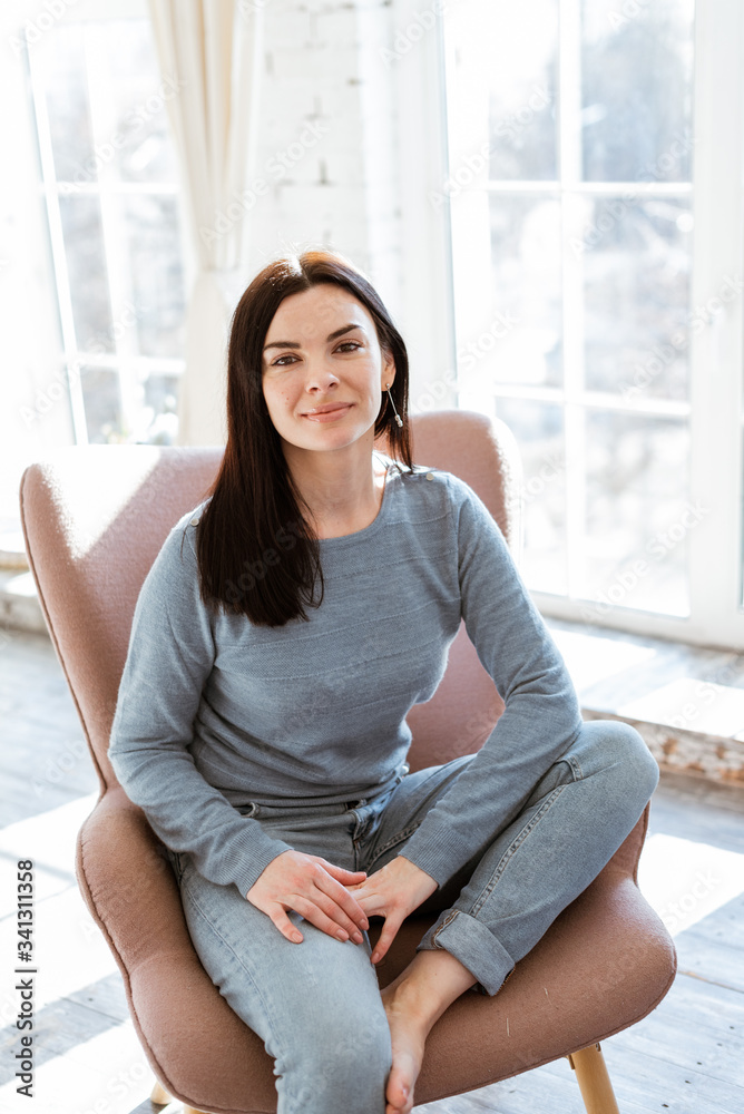 portrait of a young woman in jeans clothes sitting in a pink armchair in front of a window at home