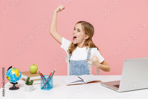 Happy little kid schoolgirl 12-13 years old study at white desk with pc laptop isolated on pink background. School distance education at home during quarantine concept. Clenching fists like winner.