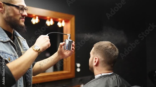 Barber sprays parfume on client in barbershop after haircut and styling. Hairdresser using old fashined sprayer with toilet water in hair salon after customer's beard shaving. Vintage cologne bottle. photo