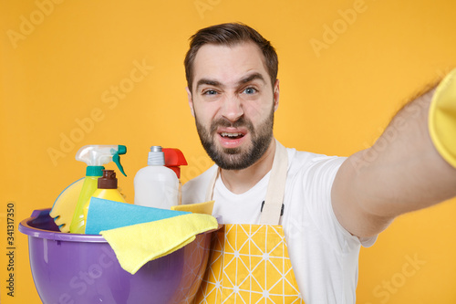 Close up of puzzled man househusband in apron hold basin detergent bottles washing cleansers doing housework isolated on yellow background. Housekeeping concept. Doing selfie shot on mobile phone. photo