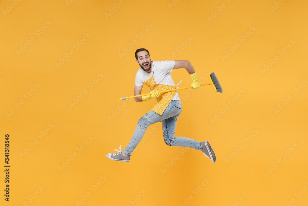 Side view of excited crazy young man househusband in apron rubber gloves hold in hands broom while doing housework isolated on yellow background studio. Housekeeping concept. Jumping, looking camera.