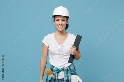 Smiling young woman in protective helmet hardhat hold clipboard with papers document isolated on blue background in studio. Instruments accessories for renovation apartment room. Repair home concept.